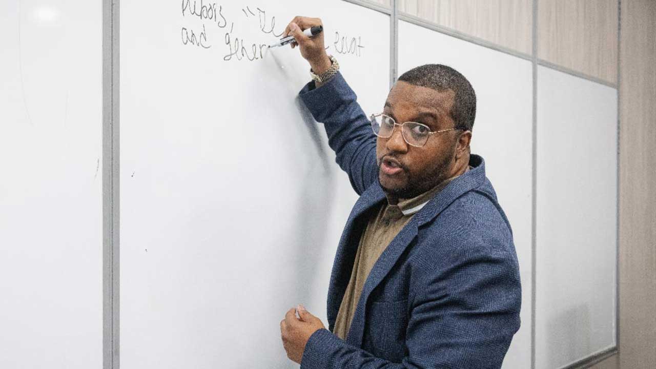 Professor Cameron Black explaining concepts on a whiteboard in a CUNY SLU classroom.