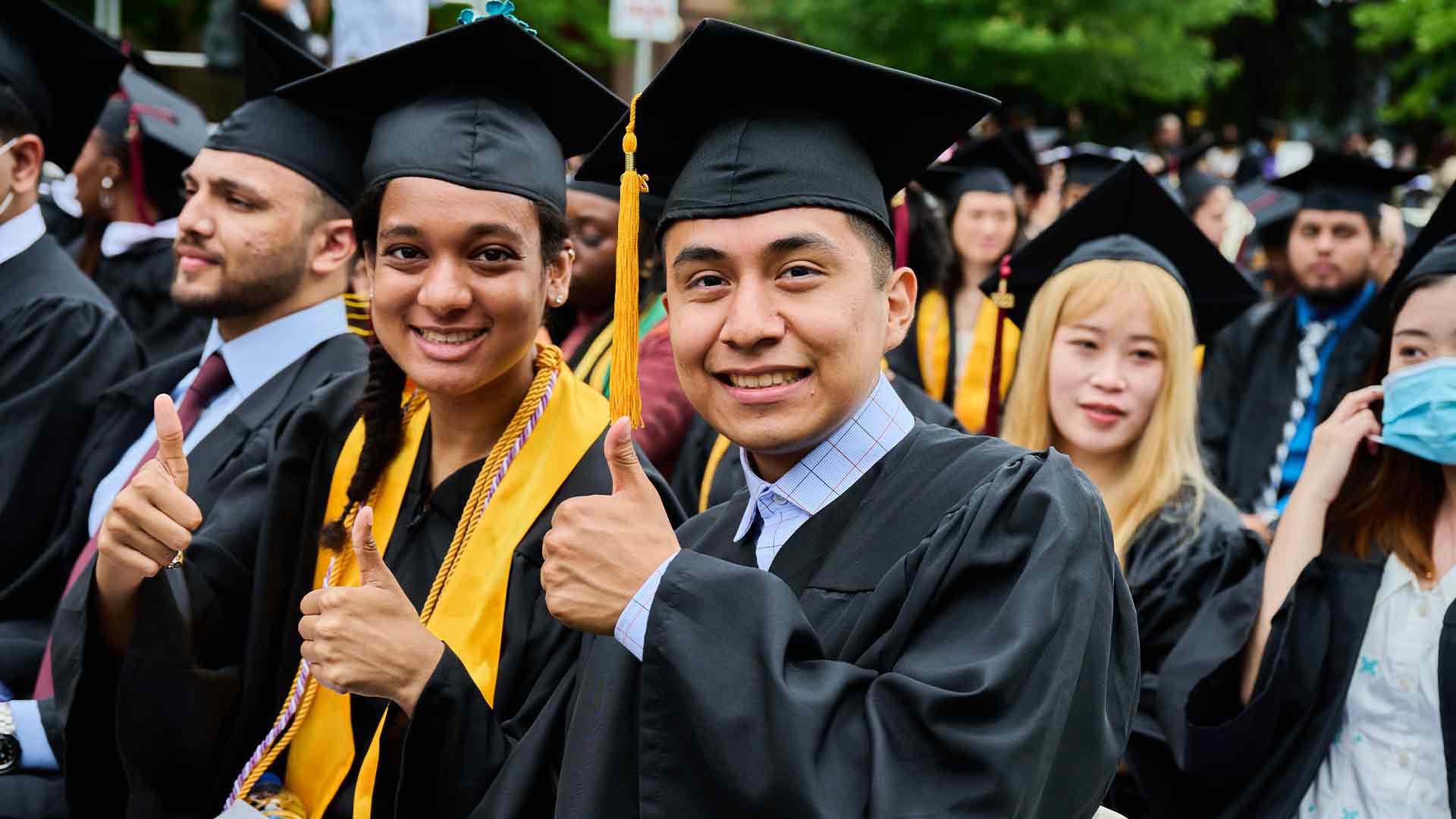 Students celebrating graduation with thumbs up at a college commencement.